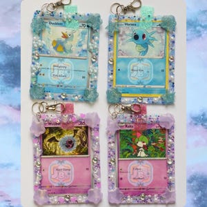 May include: Four handmade keychains featuring Pokémon trading cards. Each card is encased in a decorative frame with embellishments like pearls, glitter, and resin. The frames are in shades of blue, pink, and purple, with silver-tone clasps.