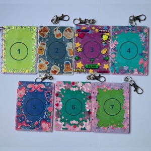 May include: A collection of seven decorative keychains. Each keychain features a rectangular design with a unique color scheme and embellishments. Some have numbers, while others showcase themed graphics like bears, hearts, and game elements. Each keychain has a silver clasp.