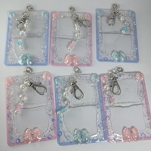 May include: A collection of six clear ID badge holders with decorative frames in pastel colors. Each holder features a bow, pearl accents, and a silver-tone clasp with a beaded chain. The frames are outlined with small, colorful embellishments.