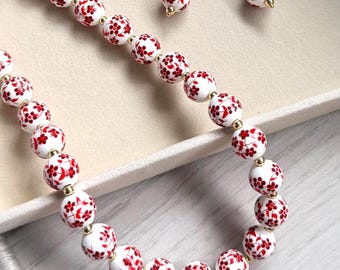 May include: A jewellery set consisting of a necklace and a pair of earrings. The necklace features white spherical beads decorated with a red floral pattern, separated by small gold-toned spacers. The earrings feature matching single beads.