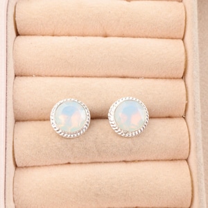 May include: Two round stud earrings with a pearlescent, opalescent center stone. The earrings have a silver-colored, rope-like border. The earrings are displayed in a pink jewelry box.