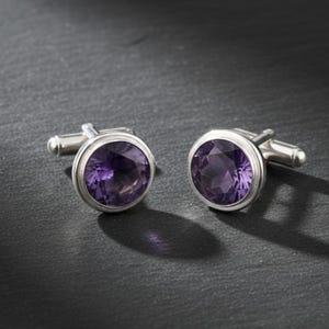 May include: A pair of silver cufflinks with a round, faceted, purple gemstone center. The cufflinks are set against a dark gray background, with the light creating a shadow.