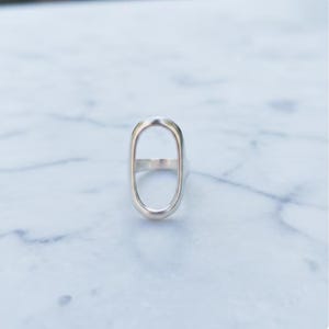 May include: A silver ring with an elongated oval design. The ring's band is thin, and the oval shape is open in the center. The ring is set against a white and gray marble background.