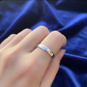 May include: A polished silver ring is worn on a finger. The ring has a smooth, rounded band and reflects light, creating a bright, shiny appearance. The background is a deep blue fabric.