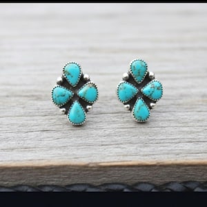 Puede incluir: Un par de pendientes de botón turquesa. Cada pendiente presenta cuatro piedras turquesas en forma de lágrima dispuestas en forma de trébol, engastadas en metal plateado. Los pendientes se muestran sobre una superficie de madera.
