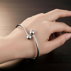 May include: A silver-colored cuff bracelet with two spherical accents. The bracelet is worn on a wrist, showcasing its simple, elegant design. The metal appears polished and reflective, creating a modern aesthetic.