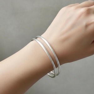 May include: Two silver-colored bangles are worn on a person's wrist. The bangles are smooth and round, reflecting light. The background is a solid, neutral gray, emphasizing the jewelry. The bangles are a simple, elegant accessory.