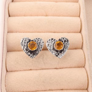 May include: A pair of heart-shaped stud earrings. The earrings have a textured silver-tone metal design with a round, orange-yellow gemstone in the center. The earrings are displayed in a pink jewelry box.