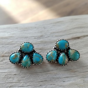 May include: A pair of turquoise cluster earrings. Each earring features five teardrop-shaped turquoise stones set in a silver-colored metal. The turquoise stones have a mix of blue and green hues.