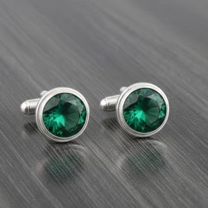 Op de afbeelding: Een paar zilveren manchetknopen met grote, ronde, smaragdgroene edelstenen. De edelstenen zijn facetgeslepen, waardoor ze het licht vangen en een schitterende glans reflecteren. Op een donkere, getextureerde achtergrond.
