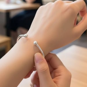May include: A silver-colored cuff bracelet with a minimalist design. The bracelet features a slender, open band that curves around the wrist, with a small, round ball at each end. The bracelet is being put on a wrist.