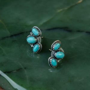 May include: A pair of silver stud earrings, each featuring three oval turquoise stones. The stones are set in a silver bezel with a decorative edge. The earrings are displayed on a green surface.