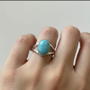 May include: A silver ring featuring a large, oval turquoise stone. The ring has a split band design, with the turquoise stone set in a bezel setting. The bright blue stone is the focal point of this piece of jewelry.