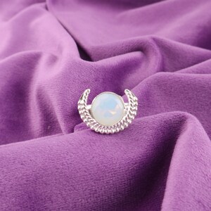 Könnte beinhalten: Silberfarbener Ring mit einem großen, runden, opalisierenden Stein in der Mitte. Das Design des Rings zeigt eine Halbmondform mit strukturierten Details auf beiden Seiten des Steins, vor einem violetten Samthintergrund.