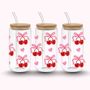 Pu&ograve; includere: Tre bicchieri di vetro trasparente con coperchi in bamb&ugrave; e cannucce di vetro. Ogni bicchiere presenta un design di ciliegie rosse legate con un fiocco rosa e piccoli cuori rosa. Sfondo rosa chiaro.