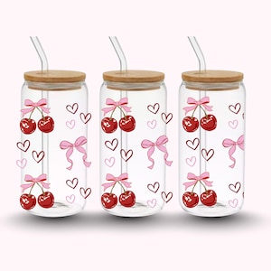 Puede incluir: Tres vasos de vidrio transparente con tapas de bambú y pajitas de vidrio. Cada vaso presenta un diseño de cerezas con lazos rosas y detalles de corazón. Las cerezas son rojas y los lazos son rosas.
