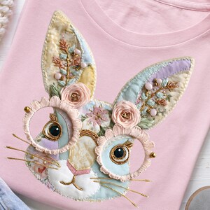 Könnte beinhalten: Hellrosa T-Shirt mit aufgesticktem Hasengesicht. Das Design zeigt ein Kaninchen mit runden Brillen und goldenen Rändern. Die Ohren sind mit Blumen und Perlen verziert, und das Kaninchen hat zarte Schnurrhaare.