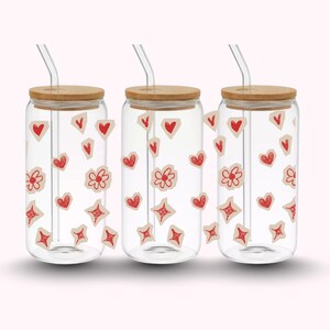 Pu&ograve; includere: Tre bicchieri di vetro trasparente con coperchi in bamb&ugrave; e cannucce di vetro. Ogni bicchiere &egrave; decorato con adesivi rossi a forma di cuore, fiore e diamante. I bicchieri sono su uno sfondo rosa chiaro.