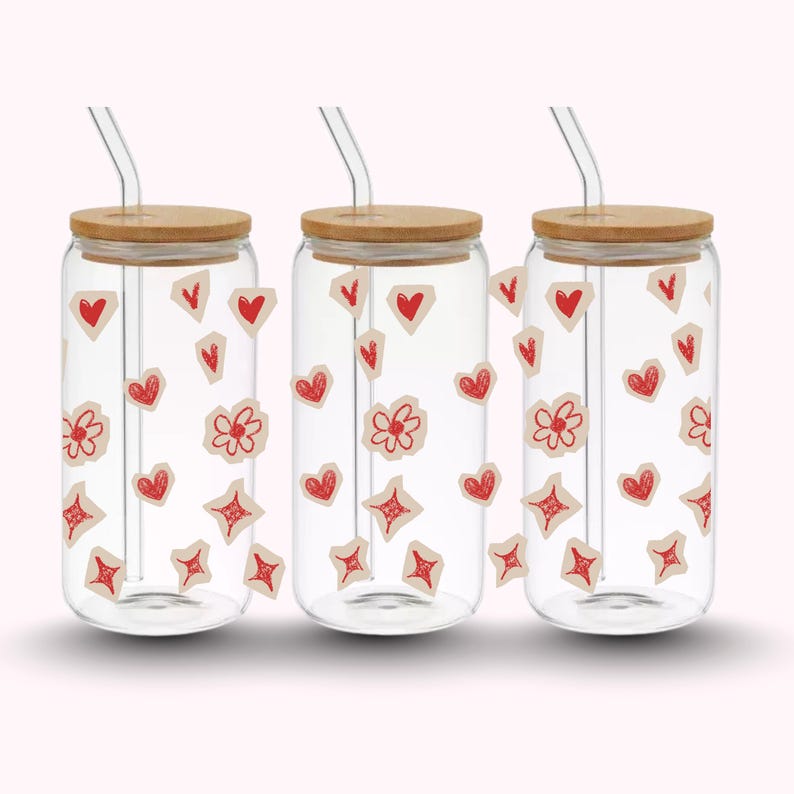Pu&ograve; includere: Tre bicchieri di vetro trasparente con coperchi in bamb&ugrave; e cannucce di vetro. Ogni bicchiere &egrave; decorato con adesivi rossi a forma di cuore, fiore e diamante. I bicchieri sono su uno sfondo rosa chiaro.