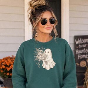 Peut inclure: Un sweat-shirt à col rond vert foncé arborant un motif central représentant un fantôme blanc tenant un grand bouquet de fleurs sauvages et de fougères. Le fantôme a deux yeux ovales noirs simples et un style de dessin au trait.