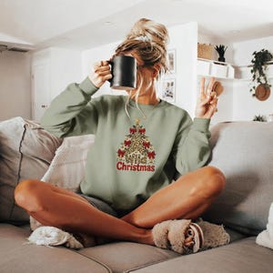 Peut inclure: Un sweat-shirt à manches longues vert sauge arborant un motif de sapin de Noël à imprimé léopard, orné de nœuds rouges et de clochettes dorées, avec l'inscription "Wild Christmas" imprimée en lettres rouges sous le motif de l'arbre.