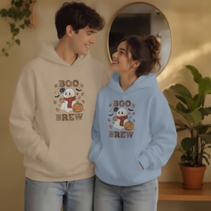 Peut inclure: Un sweat à capuche beige et un sweat à capuche bleu clair, tous deux ornés d'un motif graphique représentant un fantôme avec une écharpe rouge, tenant une tasse et à côté d'une citrouille. Le texte "BOO BREW" est imprimé en lettres marron.
