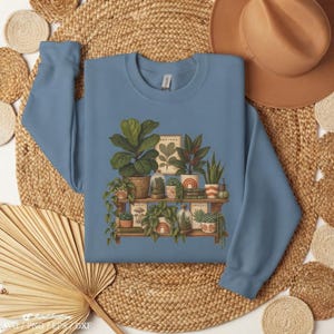 Peut inclure: Un sweat-shirt à col rond bleu poussiéreux avec une impression graphique représentant deux étagères en bois remplies de diverses plantes en pot, de petits terrariums en verre et de livres empilés. L'illustration montre un feuillage vert.