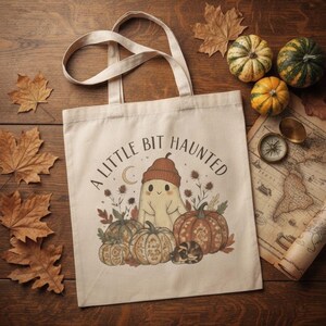 Peut inclure: Un sac fourre-tout en toile beige avec une illustration graphique d'un fantôme portant un bonnet, entouré de citrouilles et de feuillage d'automne. Le texte A LITTLE BIT HAUNTED est imprimé en arc au-dessus du fantôme avec un croissant de lune.