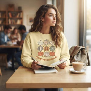 Peut inclure: Un sweat-shirt à col rond jaune pâle orné d'un motif représentant neuf citrouilles aux motifs et couleurs variés, disposées en grille. Sous les citrouilles, le texte PUMPKIN PATCH DAYDREAMS est inscrit en lettres brunes arquées.