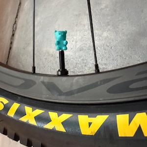 Könnte beinhalten: Eine türkisfarbene Ventilkappe in Form eines Gummibärchens auf einem schwarzen Fahrradventil. Der Reifen ist schwarz und trägt den gelben Schriftzug MAXXIS auf der Seitenwand. Das Fahrradrad hat dünne schwarze Speichen und eine dunkelgraue Felge.