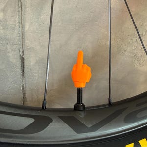 Könnte beinhalten: Eine orangefarbene Hand, die den Mittelfinger zeigt, an einer schwarzen Ventilkappe an einem Fahrradrad befestigt. Das Rad ist schwarz mit gelber Schrift auf dem Reifen. Der Hintergrund ist eine graue Betonmauer.