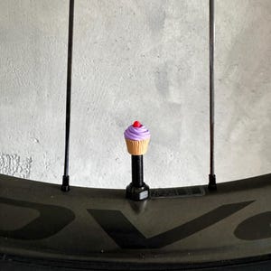 Könnte beinhalten: Eine neuartige cupcake-förmige Ventilkappe mit lila Zuckerguss und einer roten Kirsche, montiert auf einem Fahrradrad. Das Rad hat eine schwarze Felge mit gelber Schrift und schwarzen Speichen. Der Hintergrund ist eine hellgraue Wand.