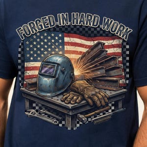 May include: A navy blue t-shirt featuring a graphic print of a welding helmet, leather gloves, and steel beams on a workbench. The design includes a United States flag background, sparks, tools, and the text FORGED IN HARD WORK in bold letters.