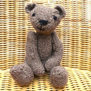 May include: A handcrafted brown teddy bear with black eyes and a black stitched nose and mouth. The bear is seated with its arms and legs extended. The bear is made of a soft, textured yarn, set against a woven wicker background.