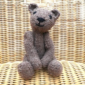 May include: A small, brown, hand-knitted teddy bear with a textured yarn surface. The bear features embroidered black thread details for the eyes, a triangular nose, and a simple curved mouth line, with rounded ears and limbs.