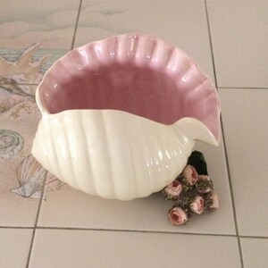May include: A white ceramic seashell planter with a pink interior. The planter is decorated with raised lines and is sitting on a tiled surface.
