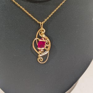 May include: A gold-colored pendant necklace featuring a vibrant red gemstone. The pendant has a wire-wrapped design with intricate swirls and curves, suspended from a delicate chain. The necklace is displayed against a dark background.
