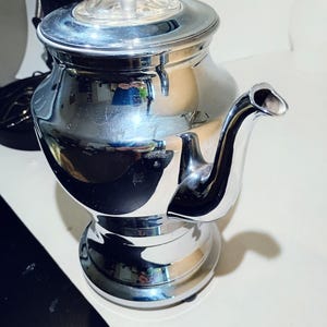 May include: A polished silver-tone metal coffee percolator with a reflective surface, a curved spout, and a clear glass knob on the lid. The vessel features a wide, rounded body that tapers toward the base and a secure, fitted lid with a glass top.
