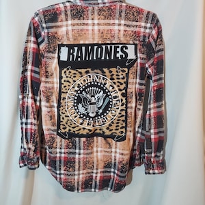 May include: A long-sleeve button-down flannel shirt featuring a red, black, and white plaid pattern with a bleached effect. The back displays a rectangular patch with the text RAMONES and a leopard print graphic featuring an eagle emblem.