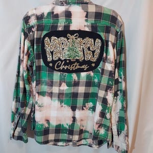 May include: A long-sleeve button-down flannel shirt in green, black, and white plaid with a bleached effect. The back features a black oval patch with the text MERRY CHRISTMAS, a leopard print font, and a green pine tree graphic.
