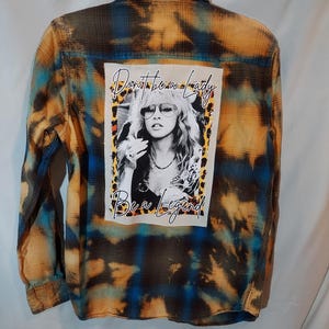 May include: A long-sleeve button-down shirt with a blue and orange bleached plaid pattern. The back features a rectangular graphic print of a woman wearing sunglasses with the text Don't be a lady, Be a legend, framed by a leopard print border.
