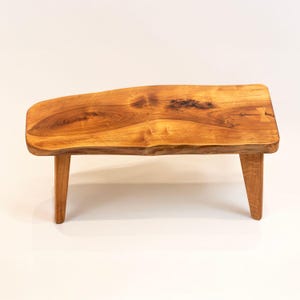 May include: A small wooden bench with a natural edge. The bench has a rectangular top with a curved edge and three angled legs. The wood has a warm, golden-brown colour with visible grain patterns. The bench is approximately 60 cm long.