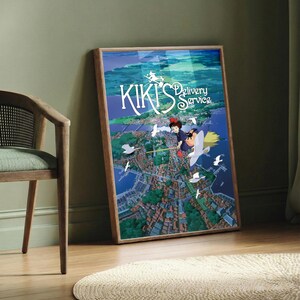 K&ouml;nnte beinhalten: Gerahmtes Poster mit dem Anime-Film "Kikis kleiner Lieferservice". Das Kunstwerk zeigt ein junges M&auml;dchen auf einem Besen, das &uuml;ber eine K&uuml;stenstadt fliegt, mit dem Titeltext in Wei&szlig;. Das Poster befindet sich in einem Holzrahmen und lehnt an einer gr&uuml;nen Wand.