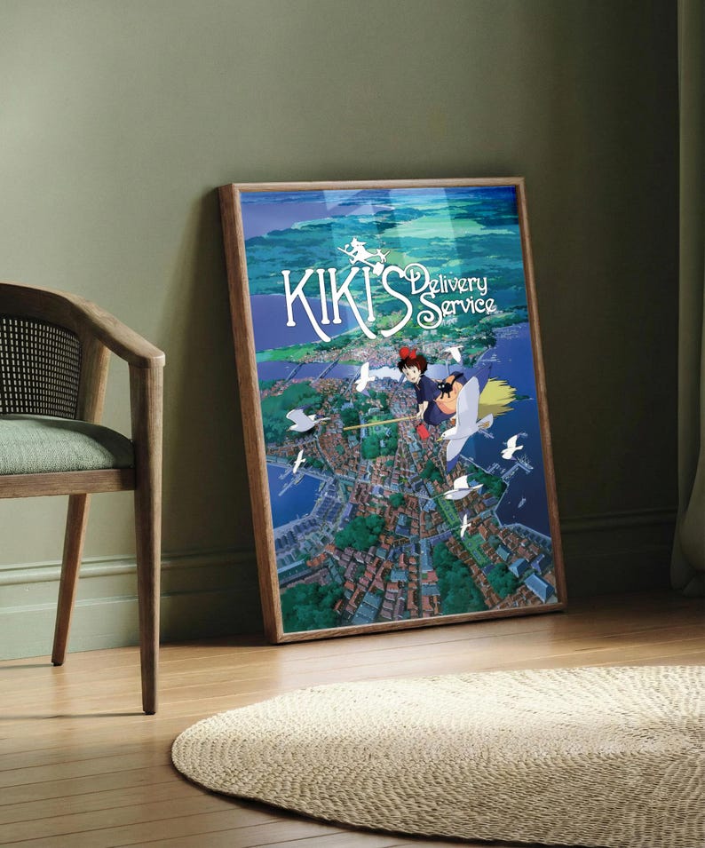 K&ouml;nnte beinhalten: Gerahmtes Poster mit dem Anime-Film "Kikis kleiner Lieferservice". Das Kunstwerk zeigt ein junges M&auml;dchen auf einem Besen, das &uuml;ber eine K&uuml;stenstadt fliegt, mit dem Titeltext in Wei&szlig;. Das Poster befindet sich in einem Holzrahmen und lehnt an einer gr&uuml;nen Wand.