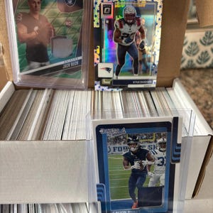 May include: A collection of football trading cards, including cards featuring Jack Bech, Kyle Dugger, and Calvin Ridley. The cards are in protective cases and are stored in a cardboard box. The cards have various designs and team logos.