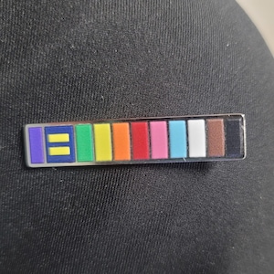 May include: A rectangular pin with a silver border featuring a series of colorful blocks. The blocks display the colors of the pride flag, including purple, yellow, green, orange, red, pink, light blue, white, brown, and black.