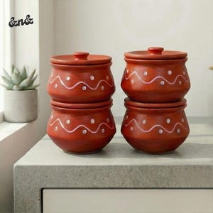 Puede incluir: Cuatro tarros de cerámica de color terracota con tapas. Cada tarro está decorado con una línea ondulada y puntos blancos. Dos tarros están apilados. Los tarros están sobre una superficie gris.