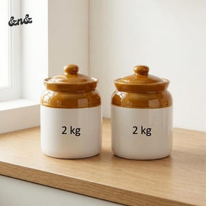 Puede incluir: Dos botes de cocina de cerámica con tapas marrones y cuerpos blancos. Cada bote tiene el texto "2 kg" impreso en la parte delantera. Los botes están sobre una superficie de madera.