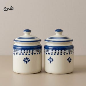 Puede incluir: Dos tarros de cerámica a juego con tapas. Los tarros son de color blanquecino con bandas decorativas azules y motivos florales. Las tapas tienen un pomo y rayas azules. Los tarros son probablemente para guardar cosas en la cocina.