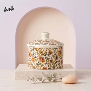 May include: A ceramic sugar bowl with a lid, featuring a floral pattern in shades of yellow, orange, and brown on a cream background. The bowl is round and sits on a square, light-colored platform.